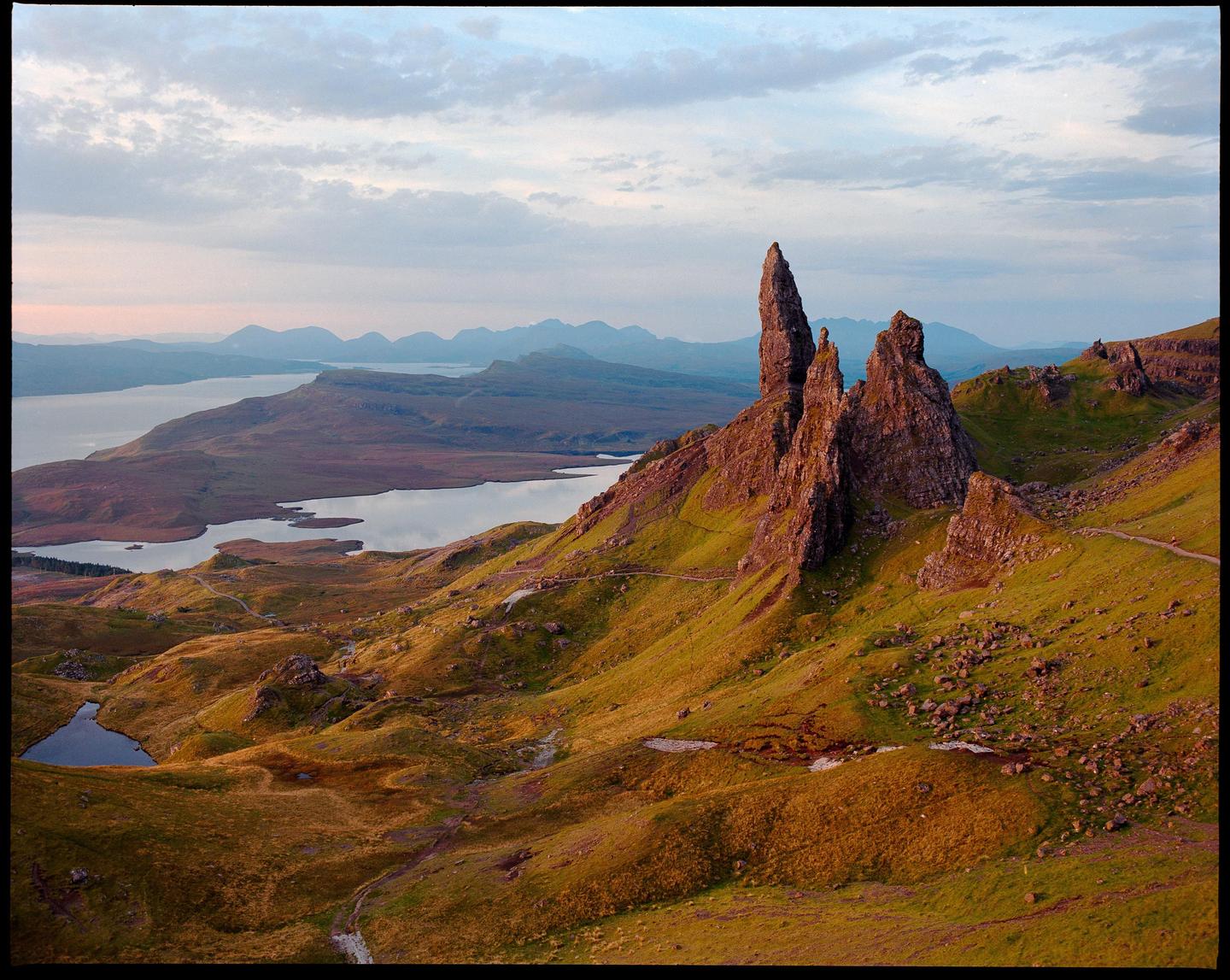 A dramatic landscape shows a large rock formation on a grassy hill overlooking a body of water and distant mountains under a cloudy sky.
