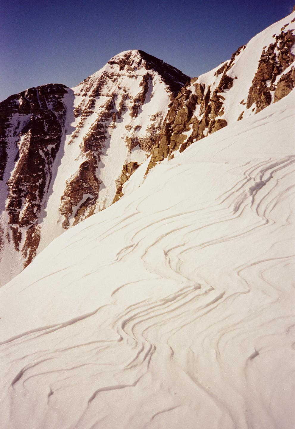 A snow-covered mountain peak rises against a clear blue sky, with textured snow in the foreground.