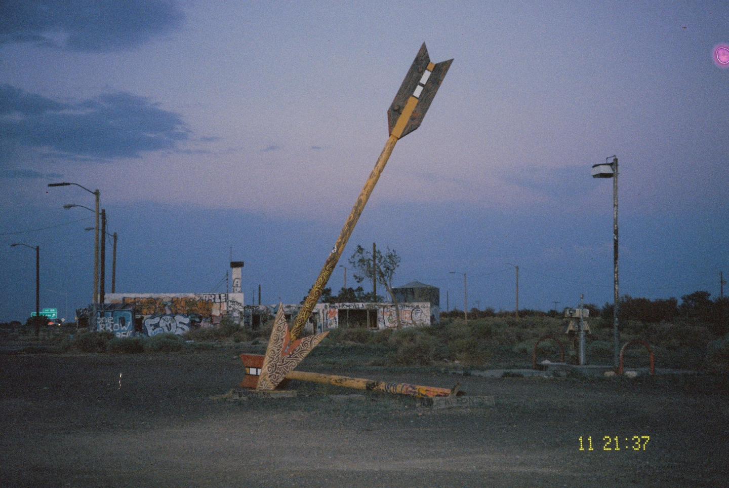 A large, broken arrow sign points towards a graffiti-covered building at dusk.