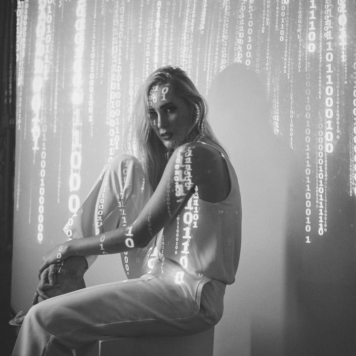 A woman sits with binary code projected onto her body and the wall behind her.