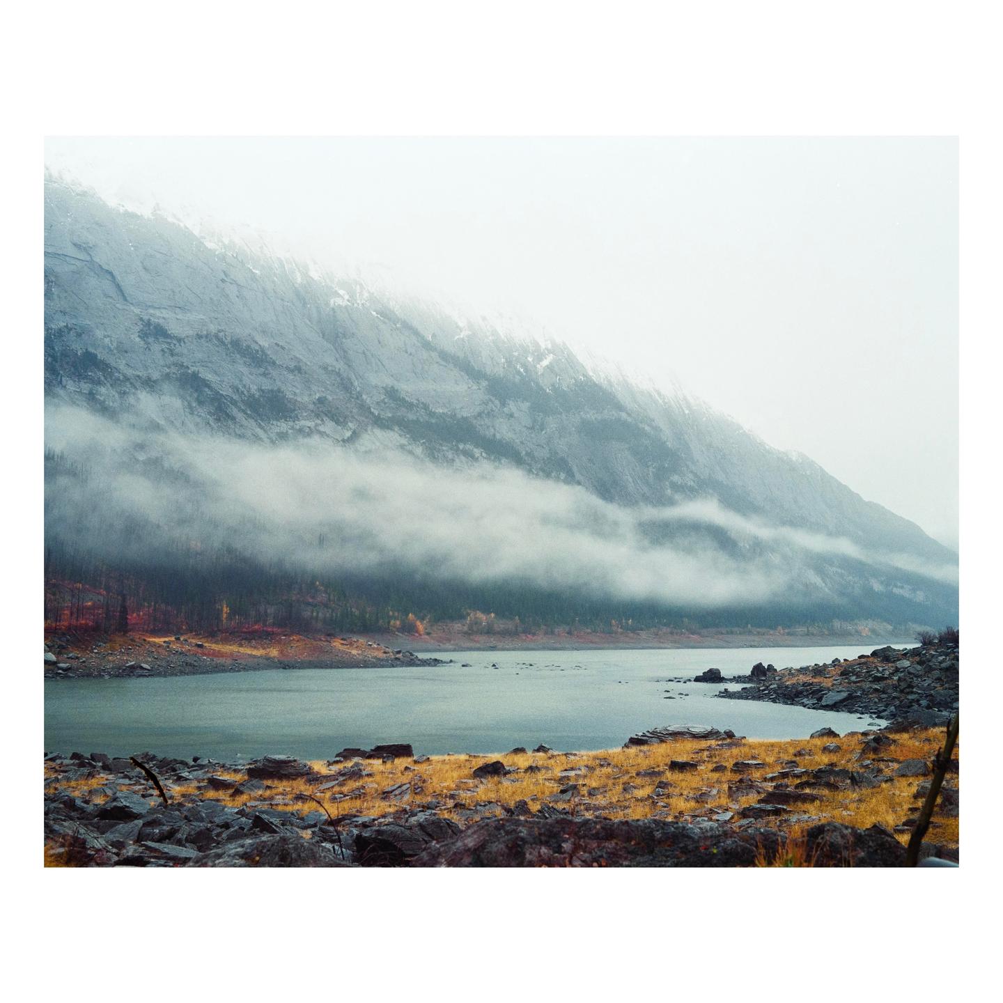 A lake is surrounded by rocky shores and mountains partially obscured by fog.