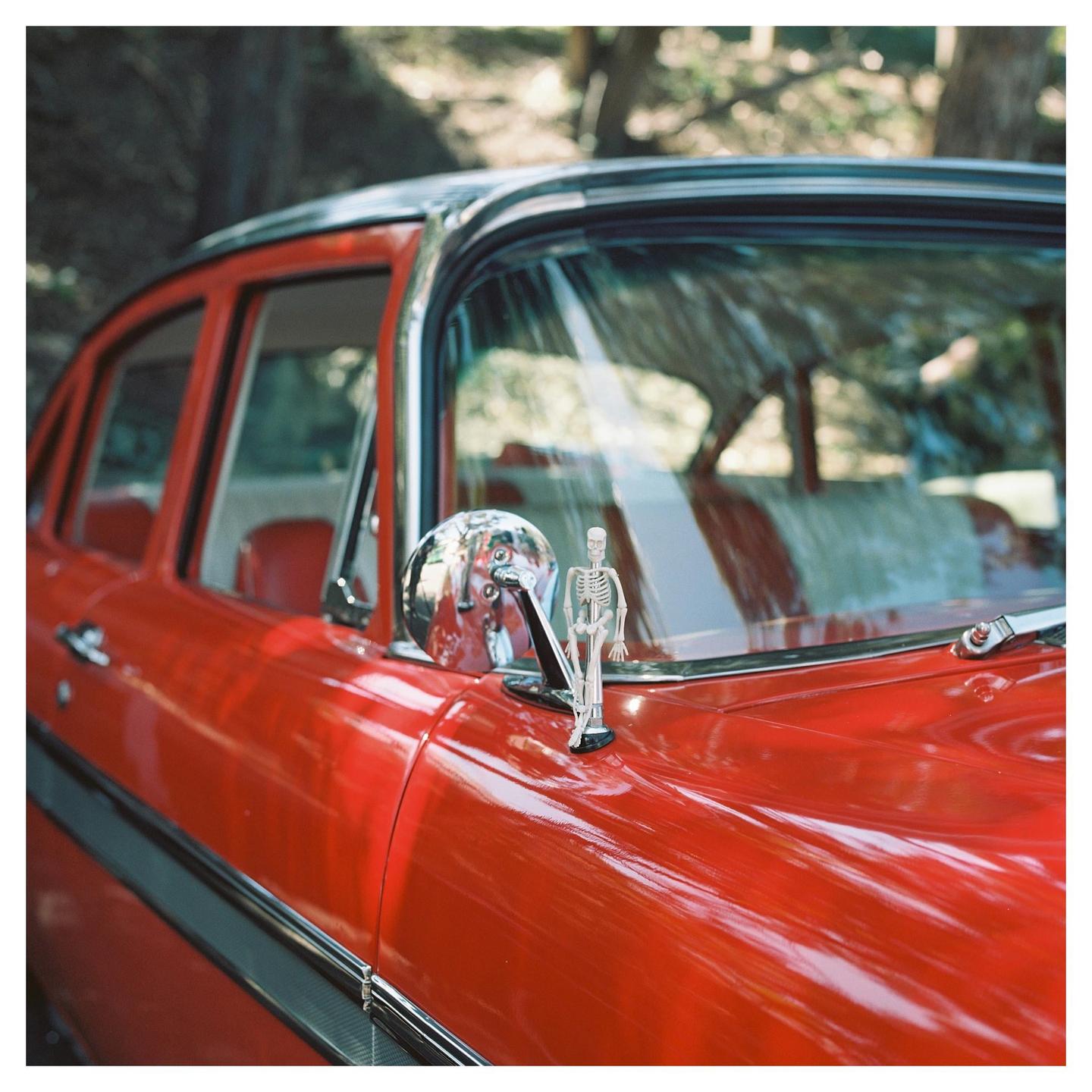 A small skeleton figurine stands on the hood of a red vintage car.
