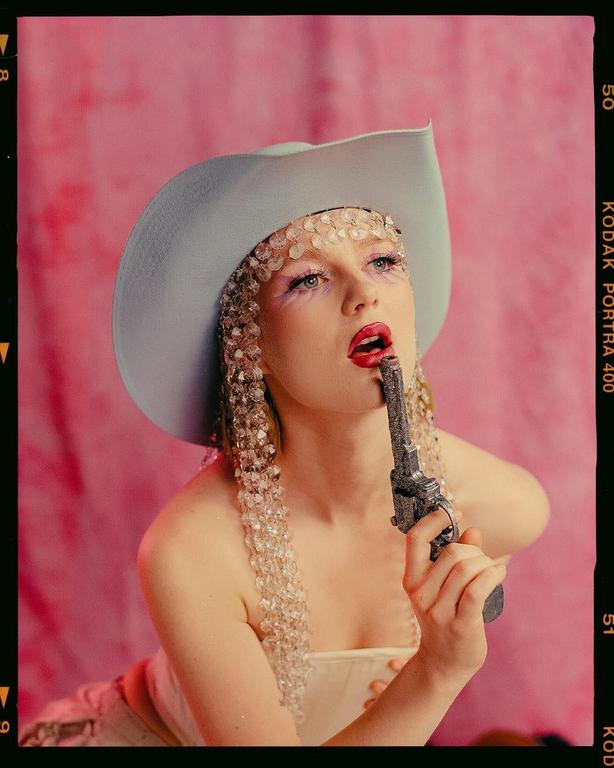 A woman wearing a cowboy hat and a beaded headpiece holds a toy gun to her mouth.