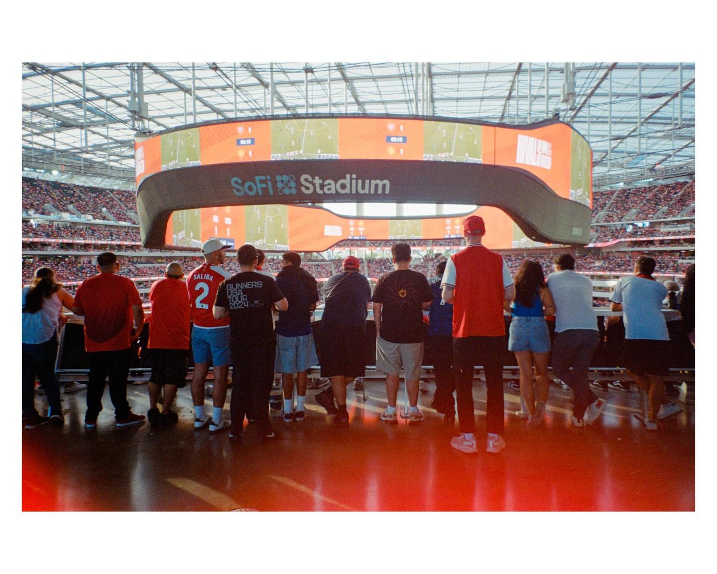 A group of people stand on a red floor looking up at a large screen inside SoFi Stadium.