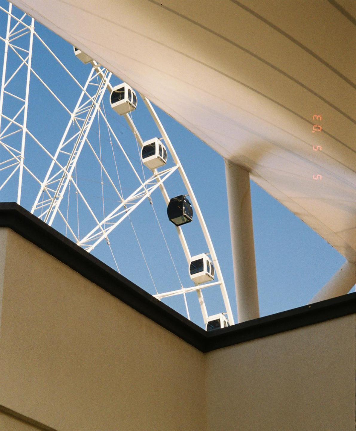 A large white ferris wheel with several gondolas is visible against a clear blue sky, framed by architectural elements.
