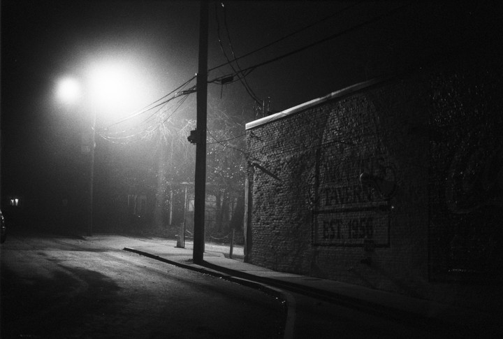 A street light illuminates a brick building with a "TAVERN EST 1956" sign on its side at night.