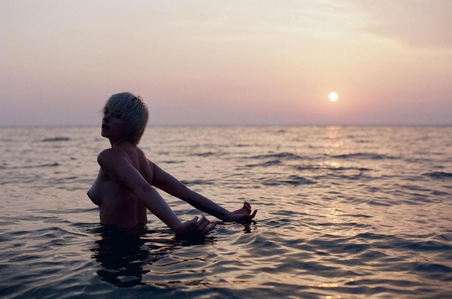 A woman with short hair is in the water with her arms outstretched as the sun sets over the ocean.