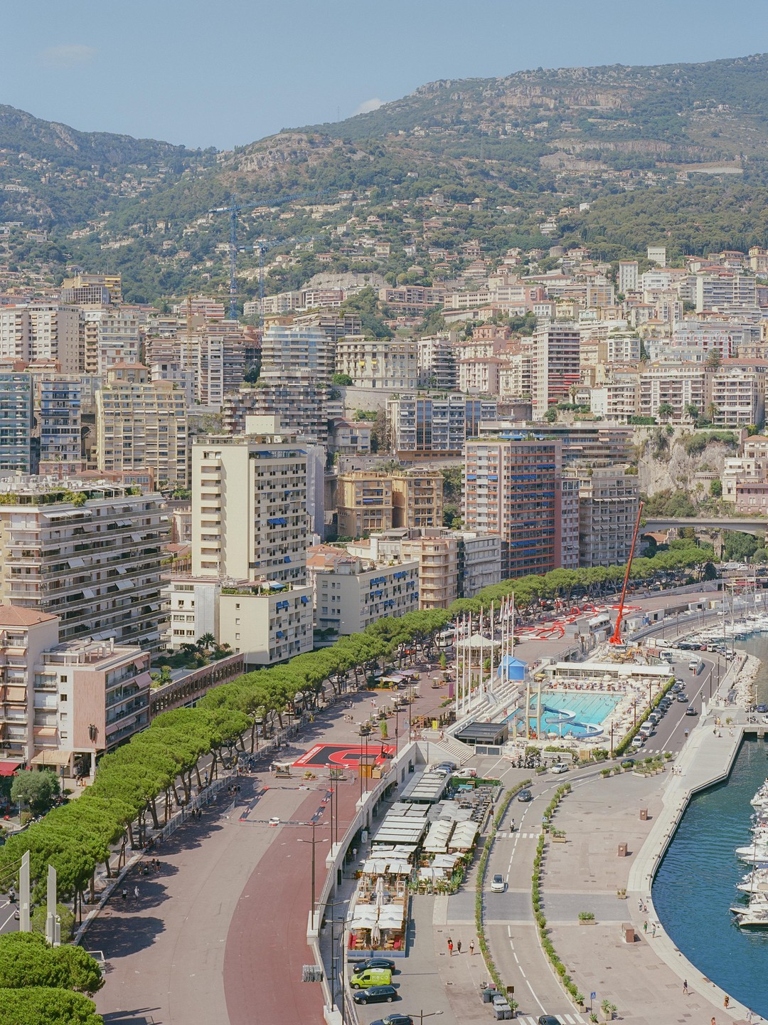 A high-angle view shows the city of Monaco with many buildings, a port with yachts, a swimming pool, and a tree-lined street, all set against a backdrop of lush green mountains.