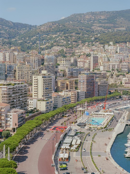 A high-angle view shows the city of Monaco with many buildings, a port with yachts, a swimming pool, and a tree-lined street, all set against a backdrop of lush green mountains.