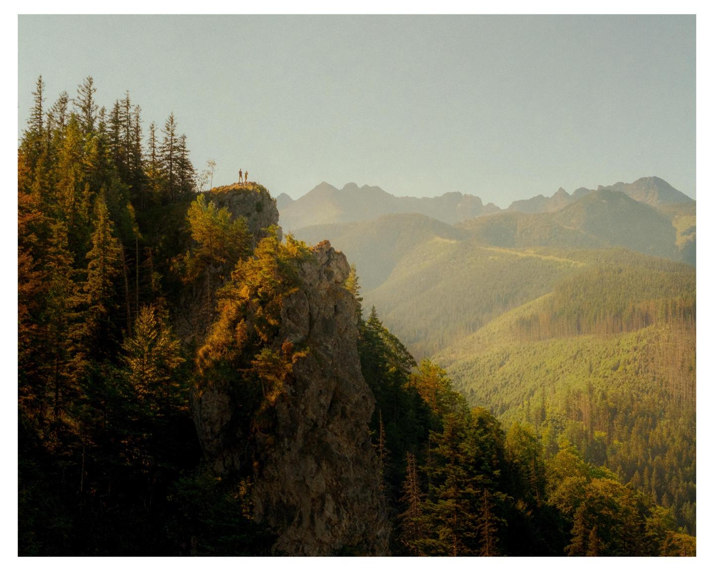Two people stand on a rocky outcrop overlooking a vast mountain landscape covered in trees.