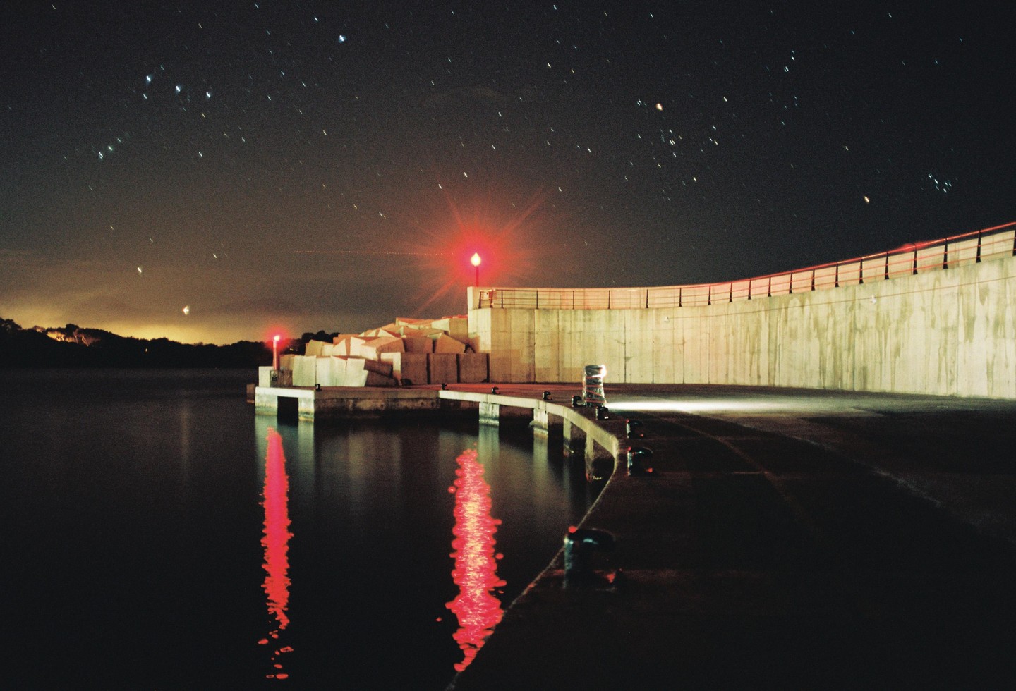 A long exposure shot of a harbor at night shows red lights reflecting on the water and stars in the sky.