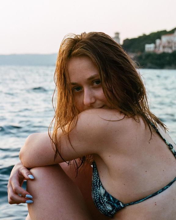 A woman with wet hair and a bikini sits by the sea, looking over her shoulder.