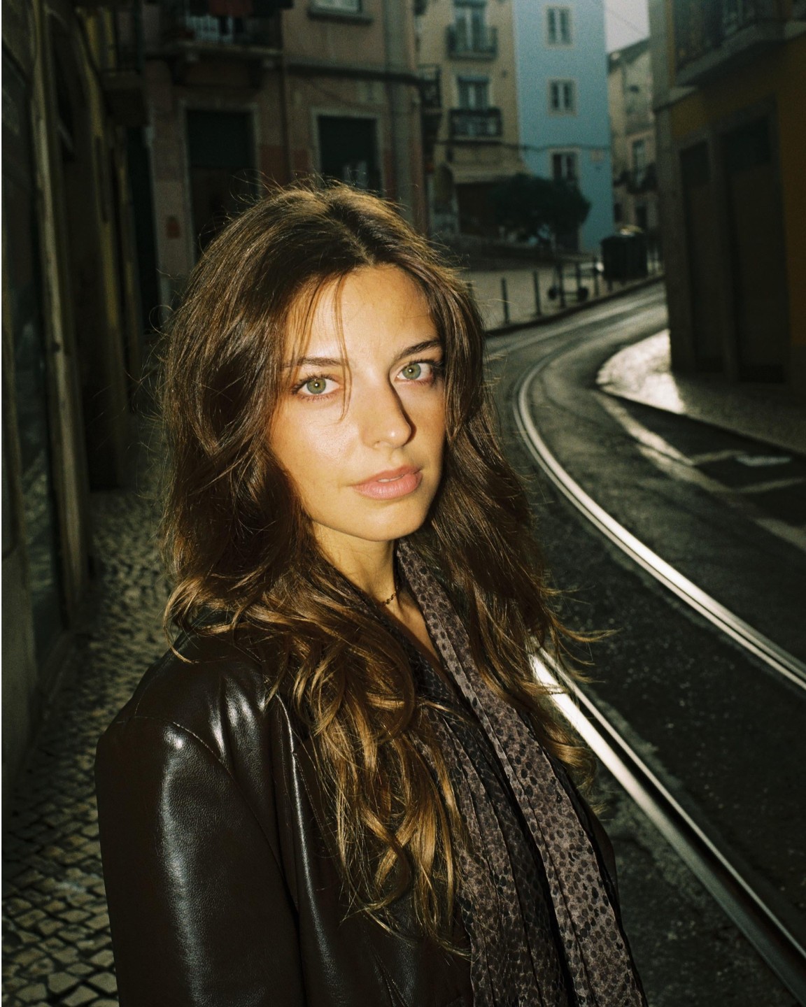 A woman with long hair and a leather jacket stands on a cobblestone street next to tram tracks.