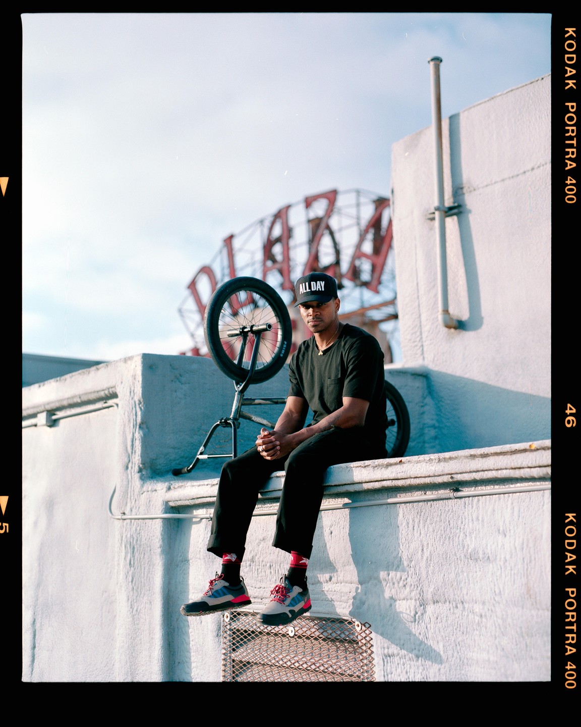 A man sits on a ledge next to a BMX bike with a large metal sign structure in the background.