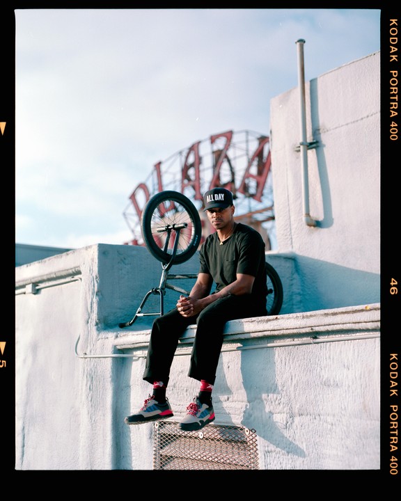 A man sits on a ledge next to a BMX bike with a large metal sign structure in the background.