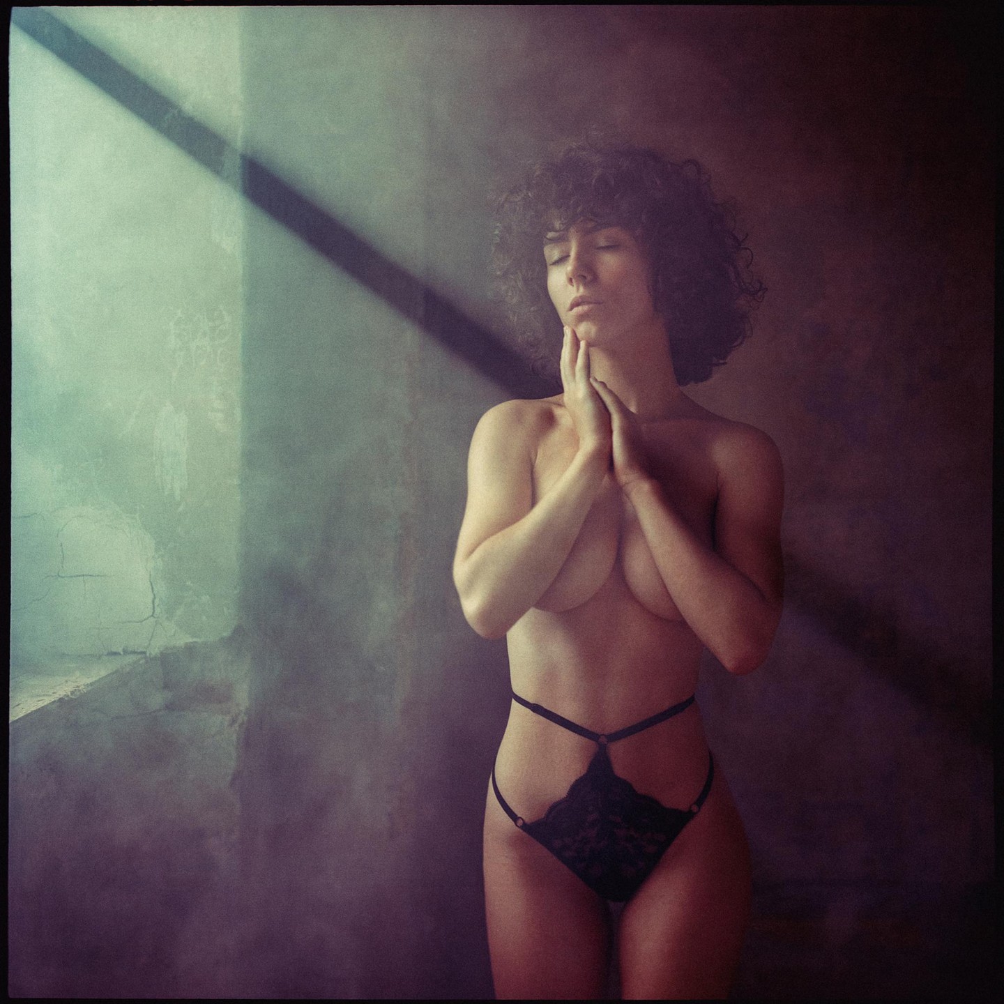 A woman with curly hair stands with her eyes closed and hands clasped under her chin, illuminated by a diagonal beam of light.