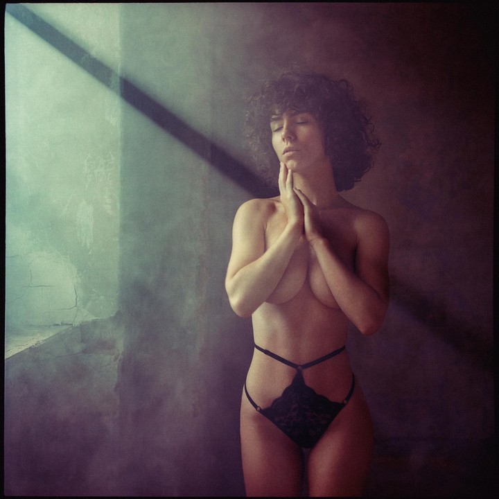 A woman with curly hair stands with her eyes closed and hands clasped under her chin, illuminated by a diagonal beam of light.