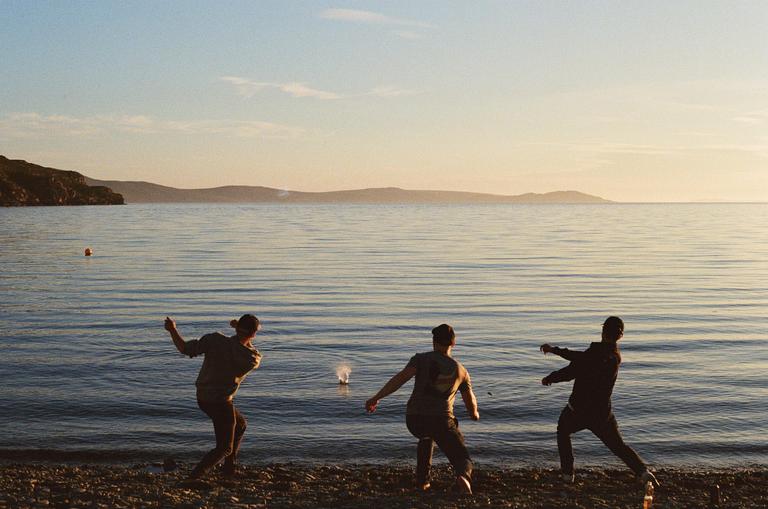 Three men are skipping rocks on a lake at sunset.