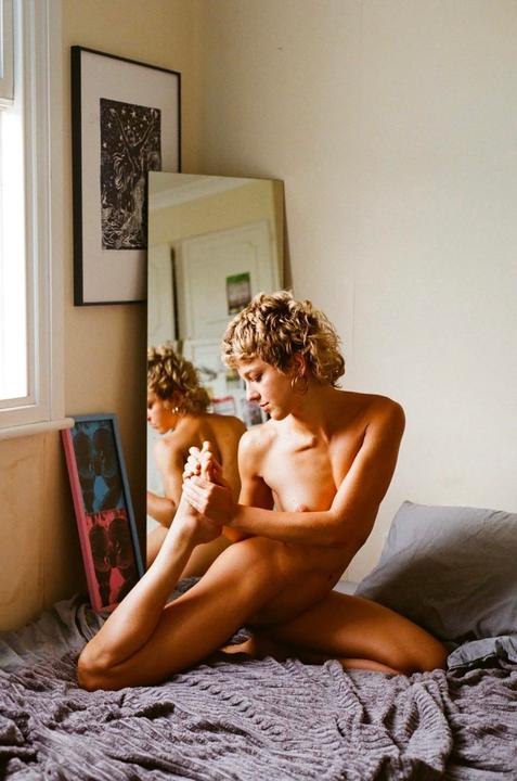 A woman with short curly hair is sitting on a bed, holding her foot, with her reflection visible in a mirror behind her.