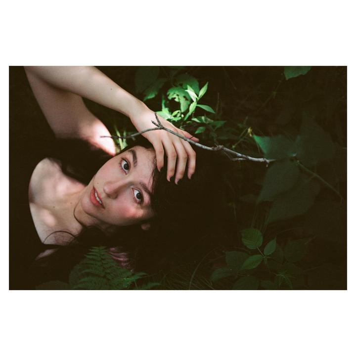 A woman with dark hair lies in a forest with her hand resting on a branch.