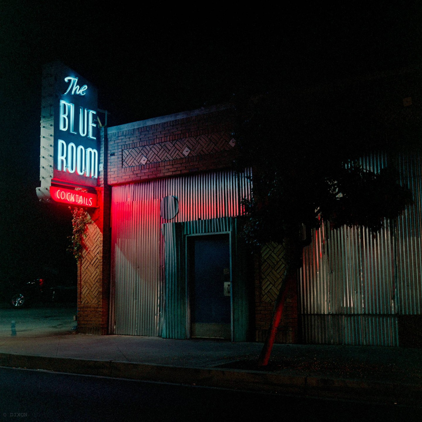 A neon sign for "The Blue Room Cocktails" illuminates a building with a corrugated metal facade at night.