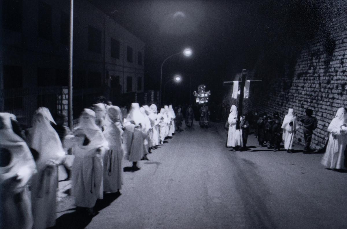 A procession of people in white robes and hoods walk down a street at night, with some carrying a large cross and another a decorated float.