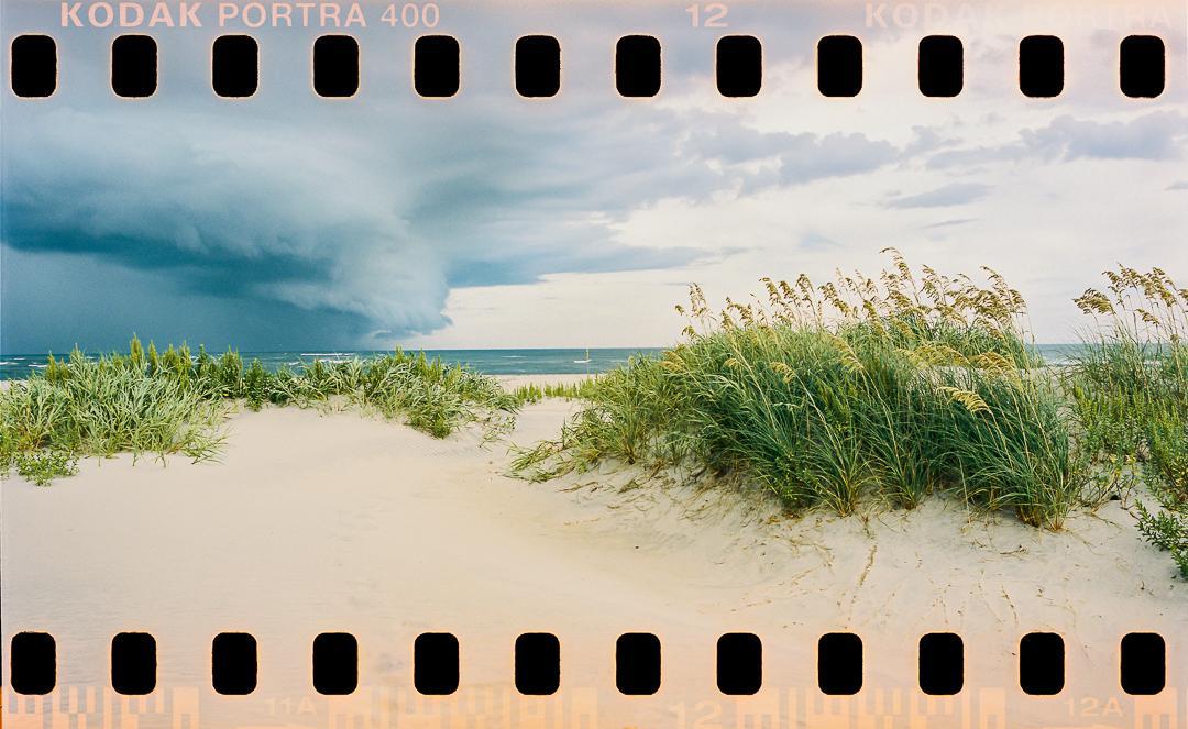 A shelf cloud looms over a sandy beach with sea oats and ocean waves.