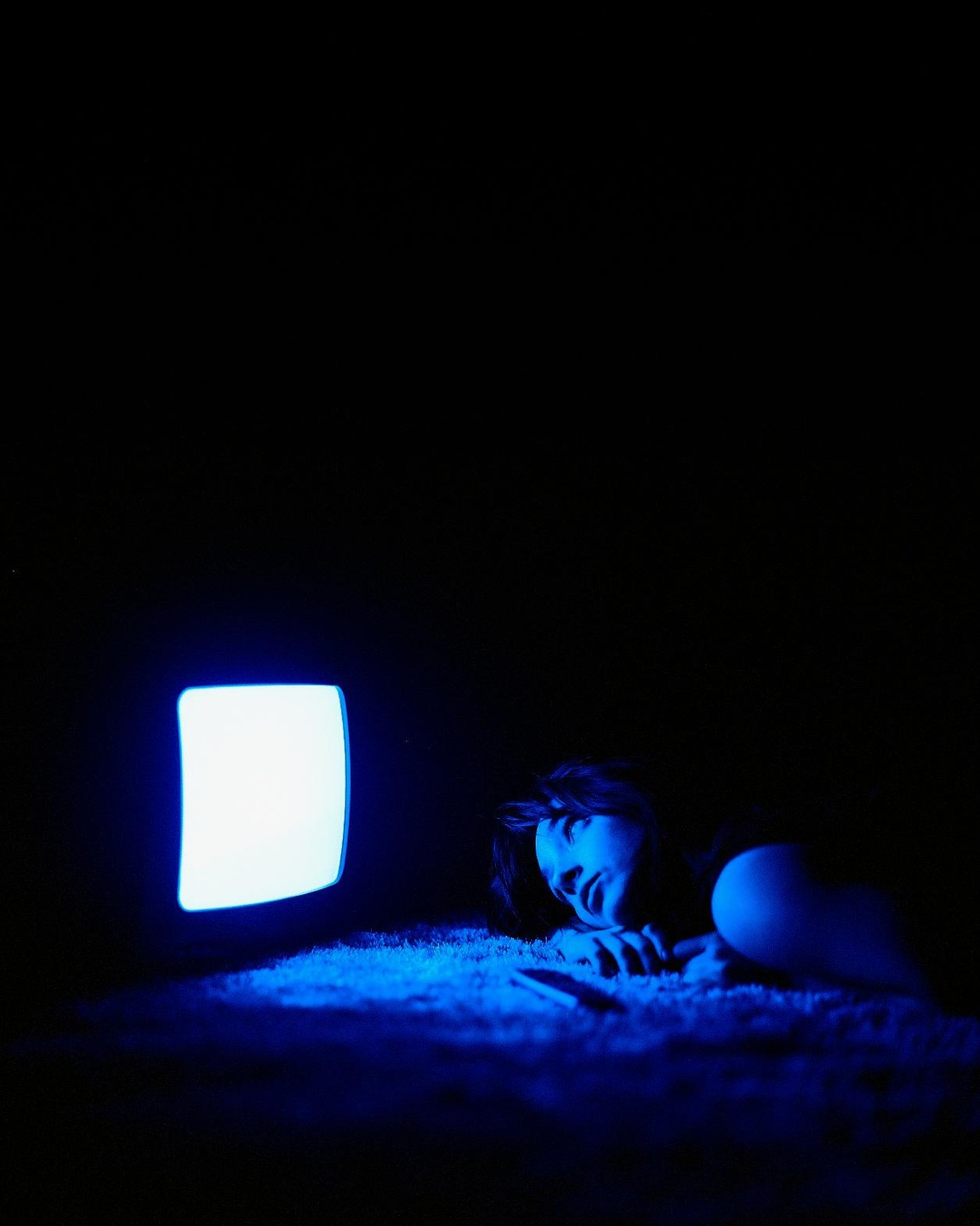 A woman lies on a carpet in a dark room, illuminated by the blue light of a television screen.