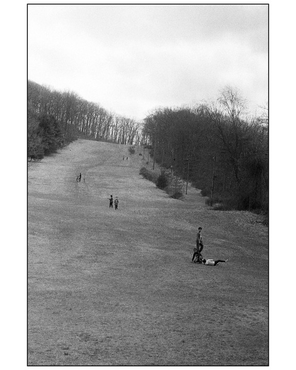 Several people are scattered across a grassy hill with trees lining both sides under an overcast sky.