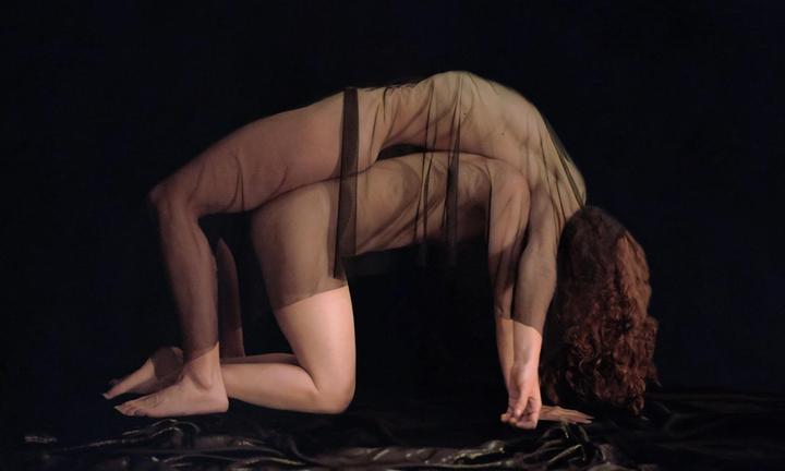 A woman in a sheer black garment is posed in a backbend on a dark surface.