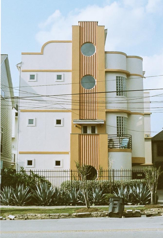 A white and yellow art deco building with round windows and a balcony is seen from across the street.