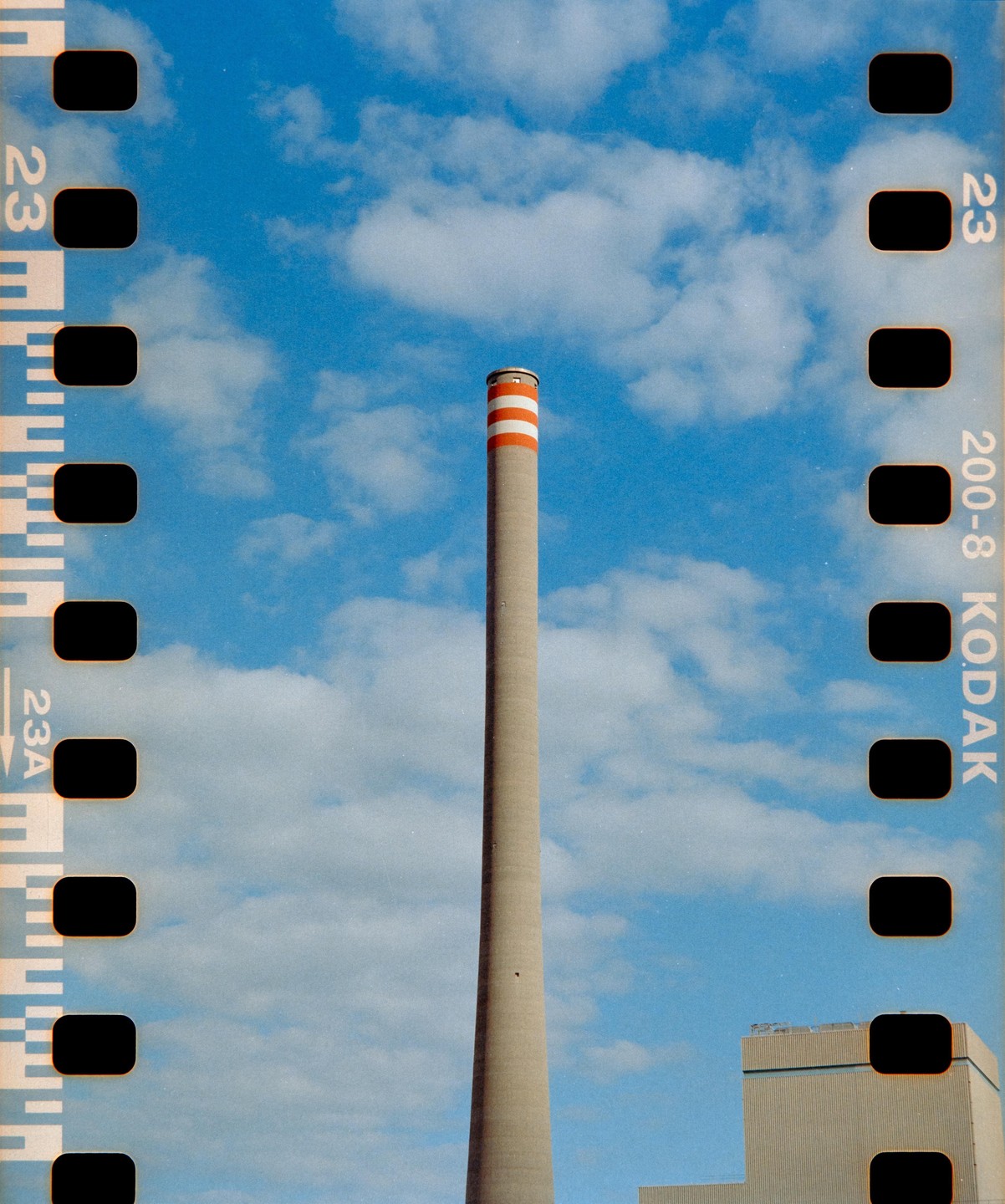 A tall, slender smokestack with red and white stripes at the top stands against a blue sky with white clouds.