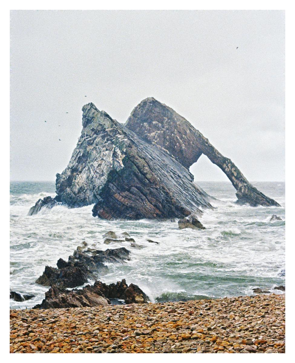 Waves crash against a rocky shore with a natural arch formation in the water.