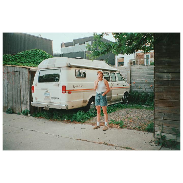 A woman stands on a sidewalk next to a parked camper van, smoking.