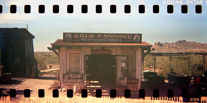 A gold panning shop with a sign that reads "Gold Panning Prospector's Palace" stands in a desert landscape with mountains in the background.