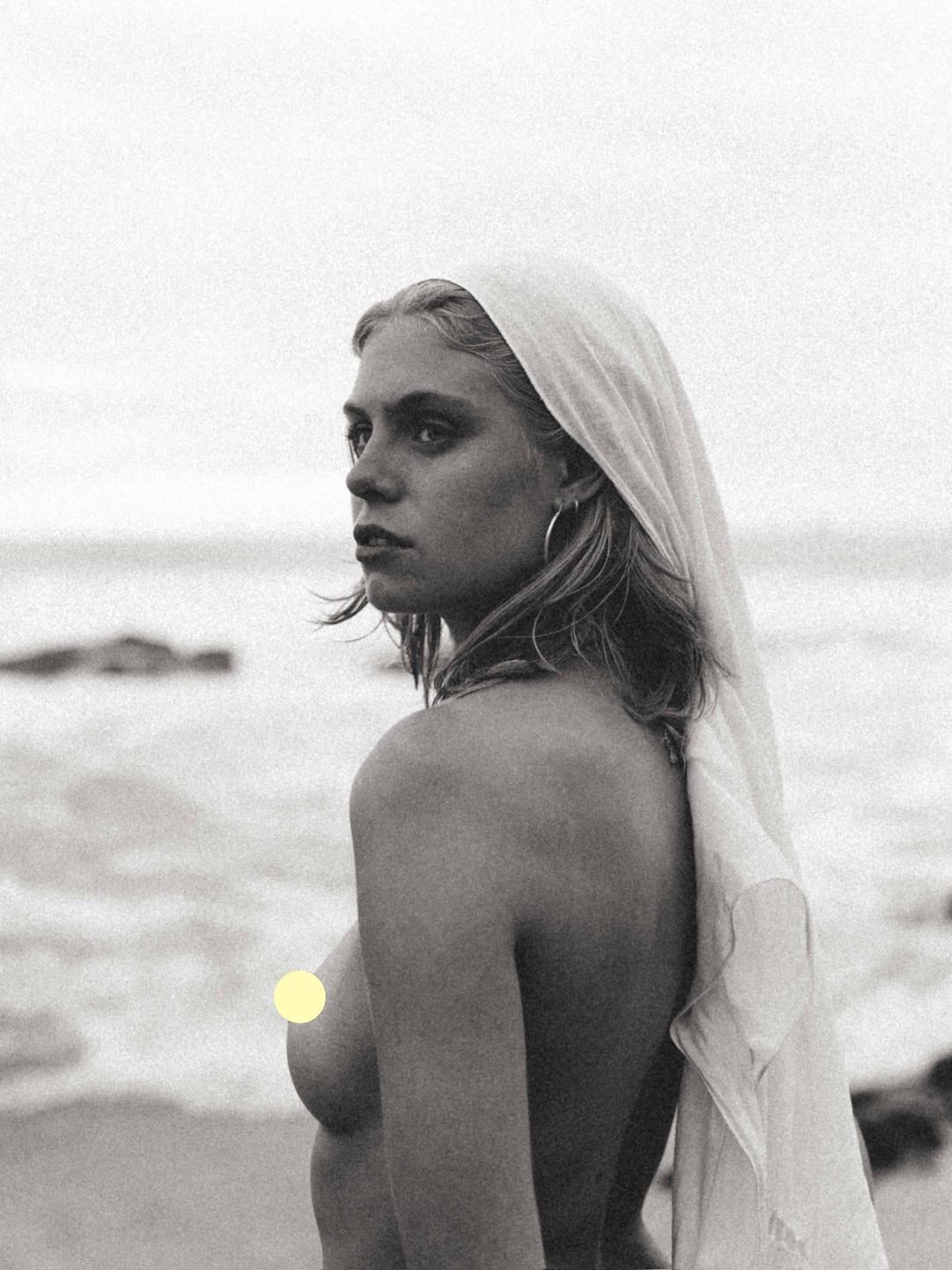 A woman with a sheer scarf over her head looks over her shoulder at the ocean.