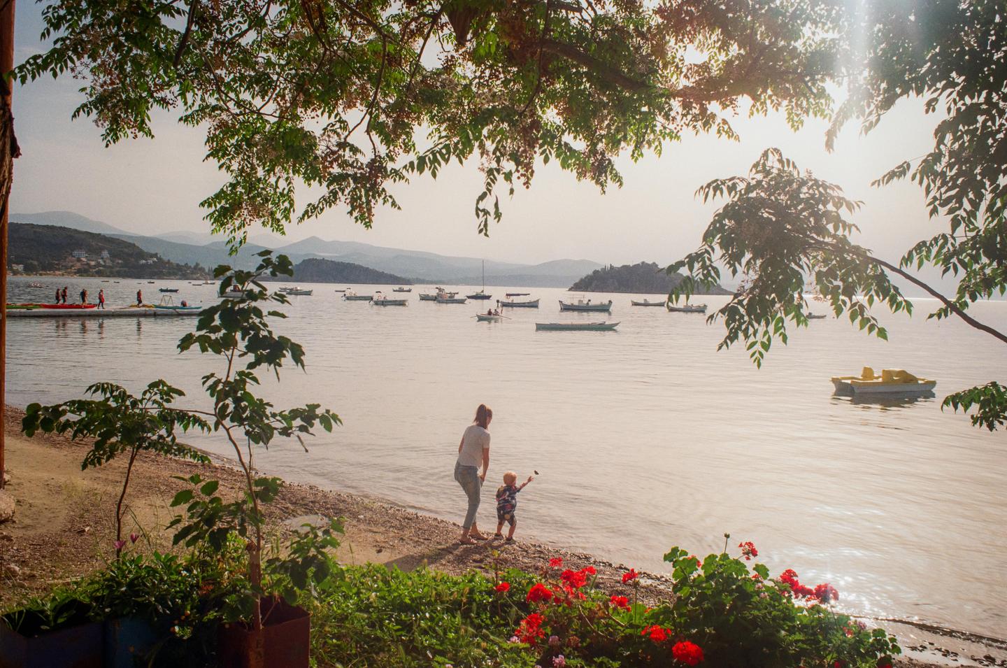 A woman and a child stand on a rocky beach next to a calm body of water with many boats, framed by green foliage.