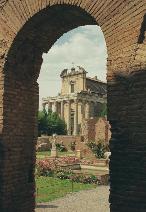 A brick archway frames a view of the Roman Forum with a classical building, ruins, a statue, and a rose garden.