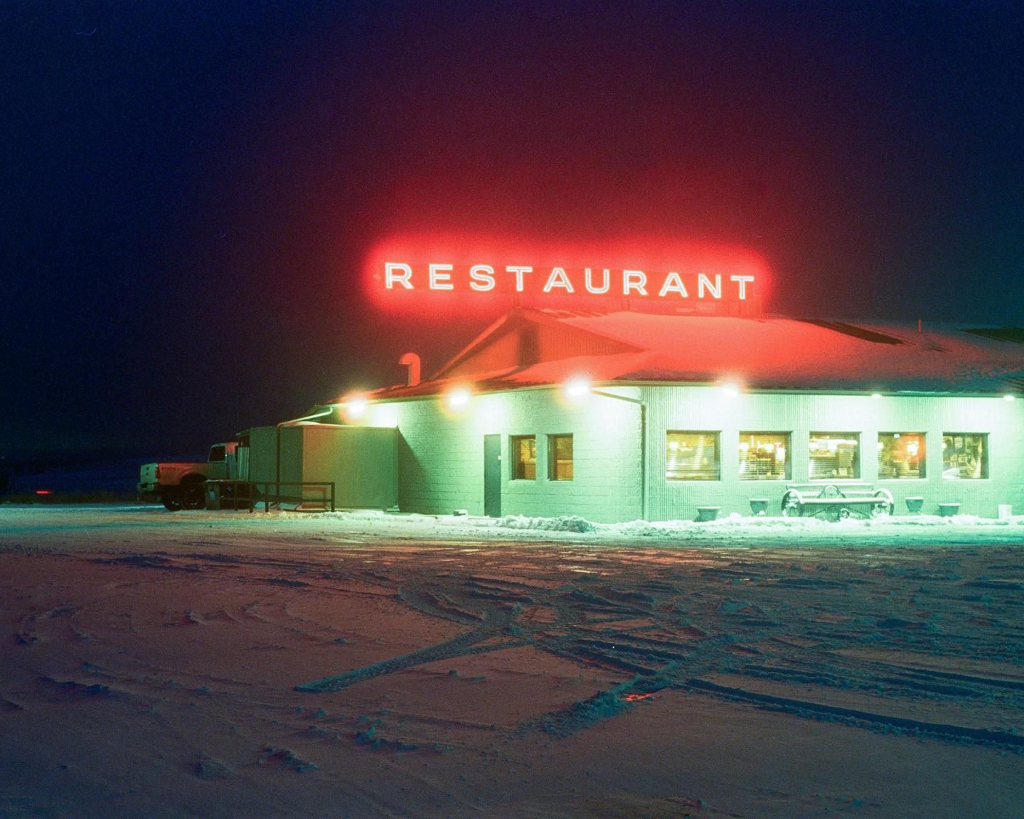 A restaurant with a red neon sign is illuminated at night, surrounded by snow.
