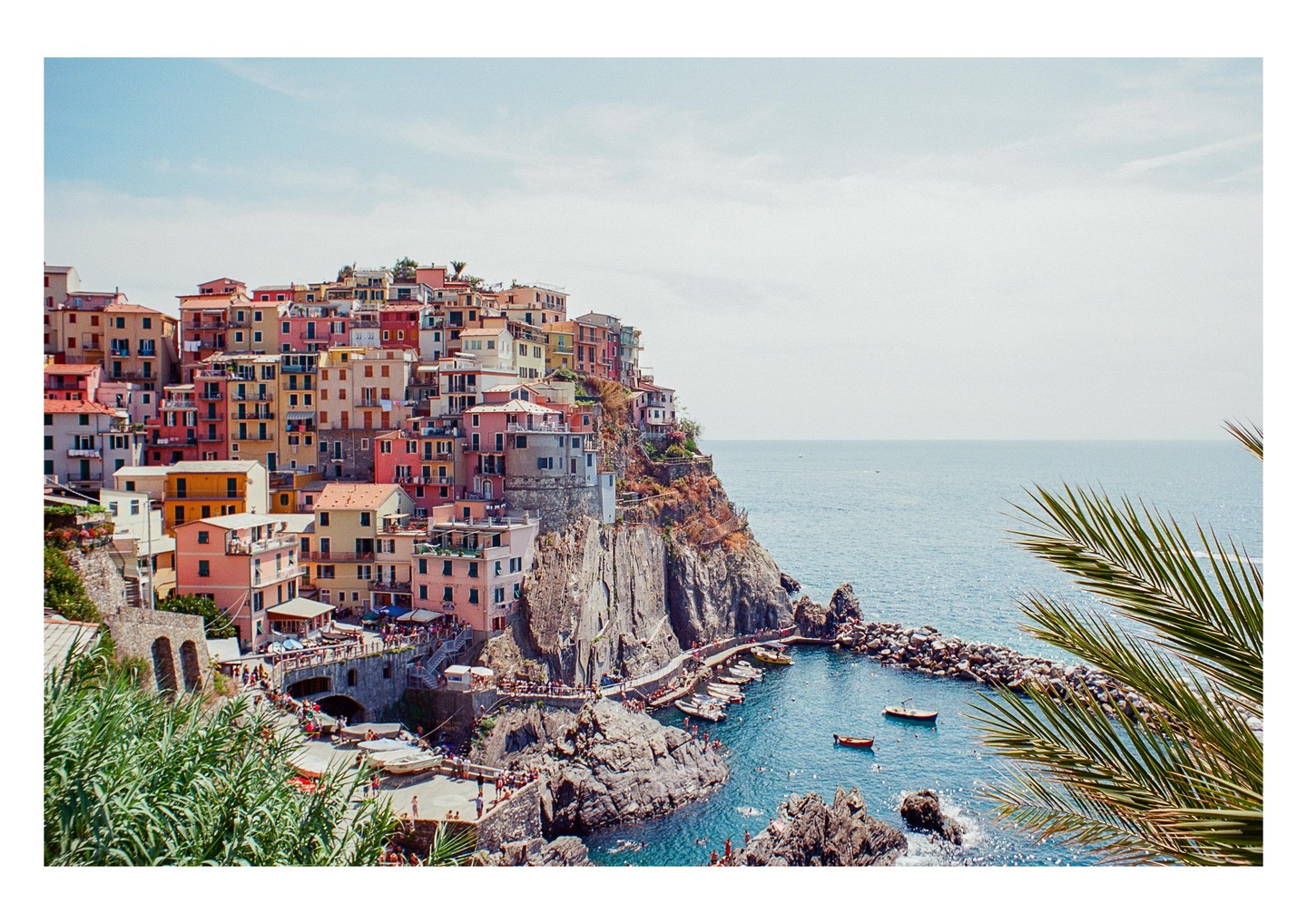 A colorful village is built into a cliffside overlooking a small harbor with boats and people swimming.