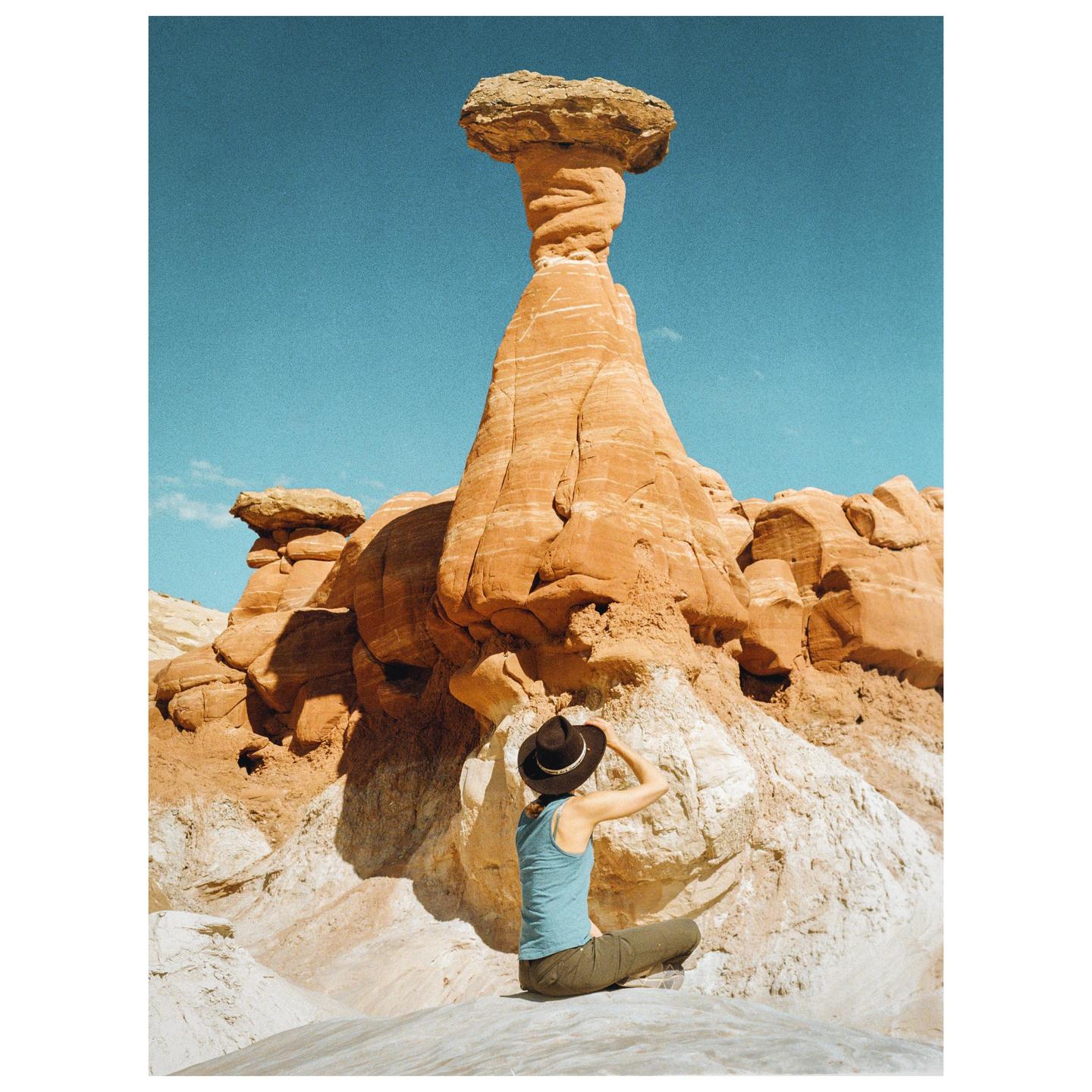 A woman sits on a rock formation looking up at a tall hoodoo under a clear blue sky.
