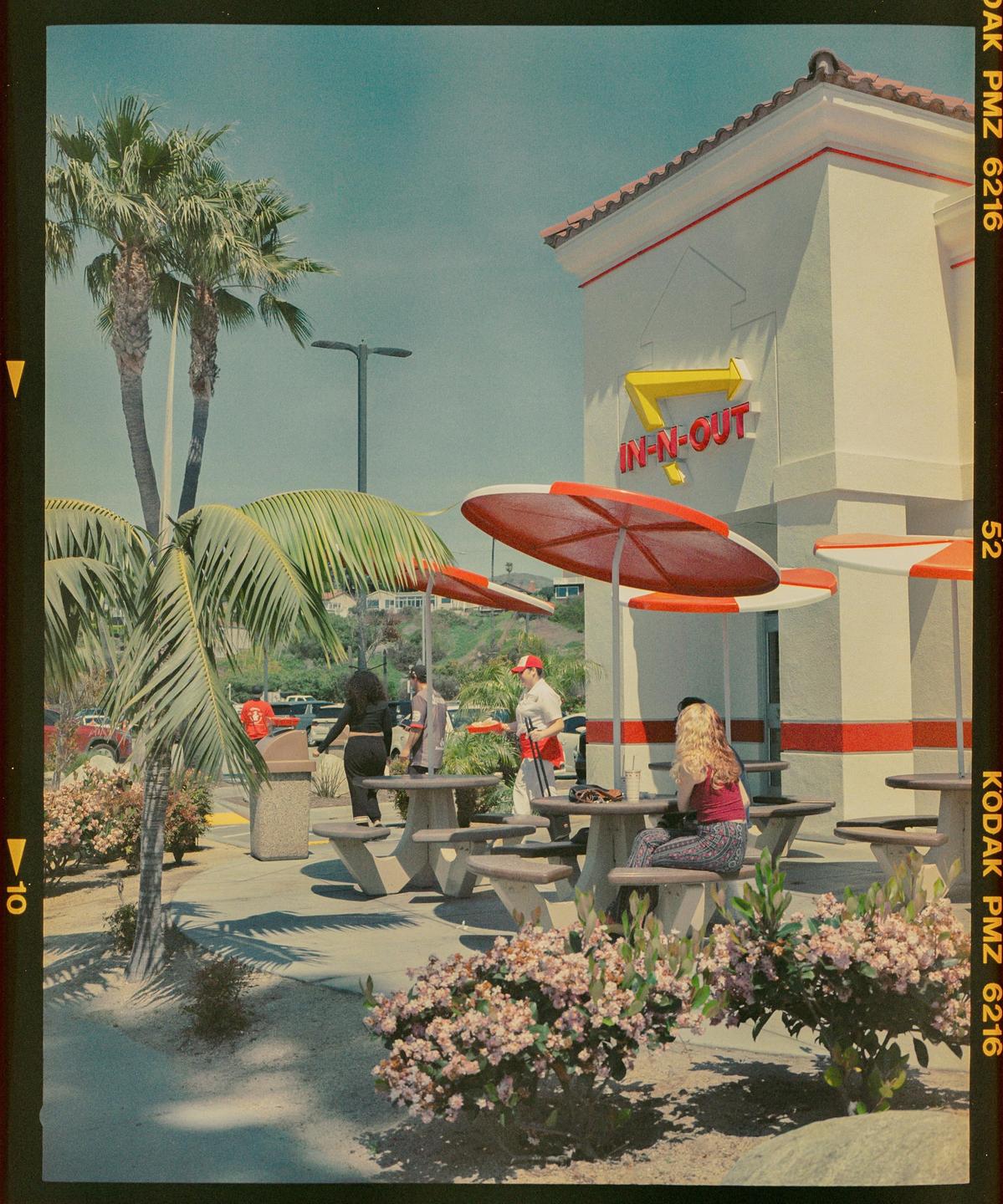 People are gathered outside an In-N-Out Burger restaurant with palm trees and flowering bushes.
