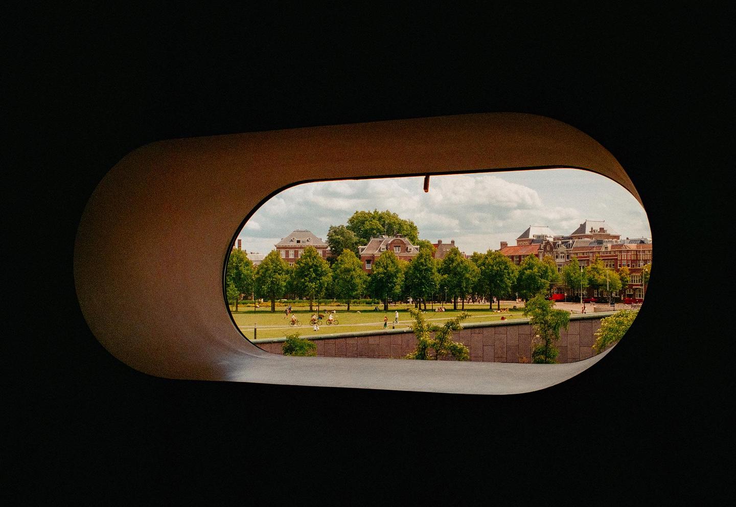 A park with trees and buildings is visible through an oval-shaped opening in a dark wall.