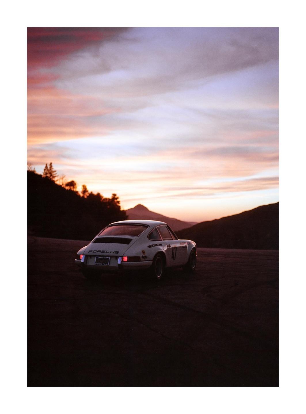 A white Porsche car is parked on a dirt road with mountains and a colorful sunset in the background.