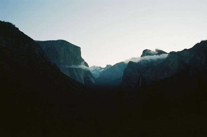 A wide valley is surrounded by tall, dark mountains with clouds clinging to their peaks and a waterfall visible on the right.