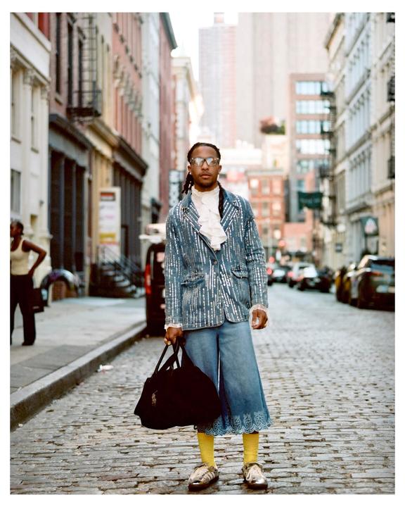 A man wearing a sequined jacket, culottes, and yellow socks stands on a cobblestone street holding a bag.
