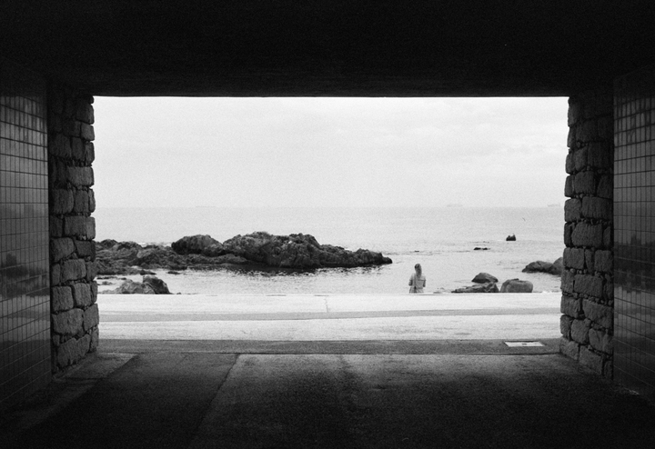 A woman stands by the ocean, framed by a dark tunnel.