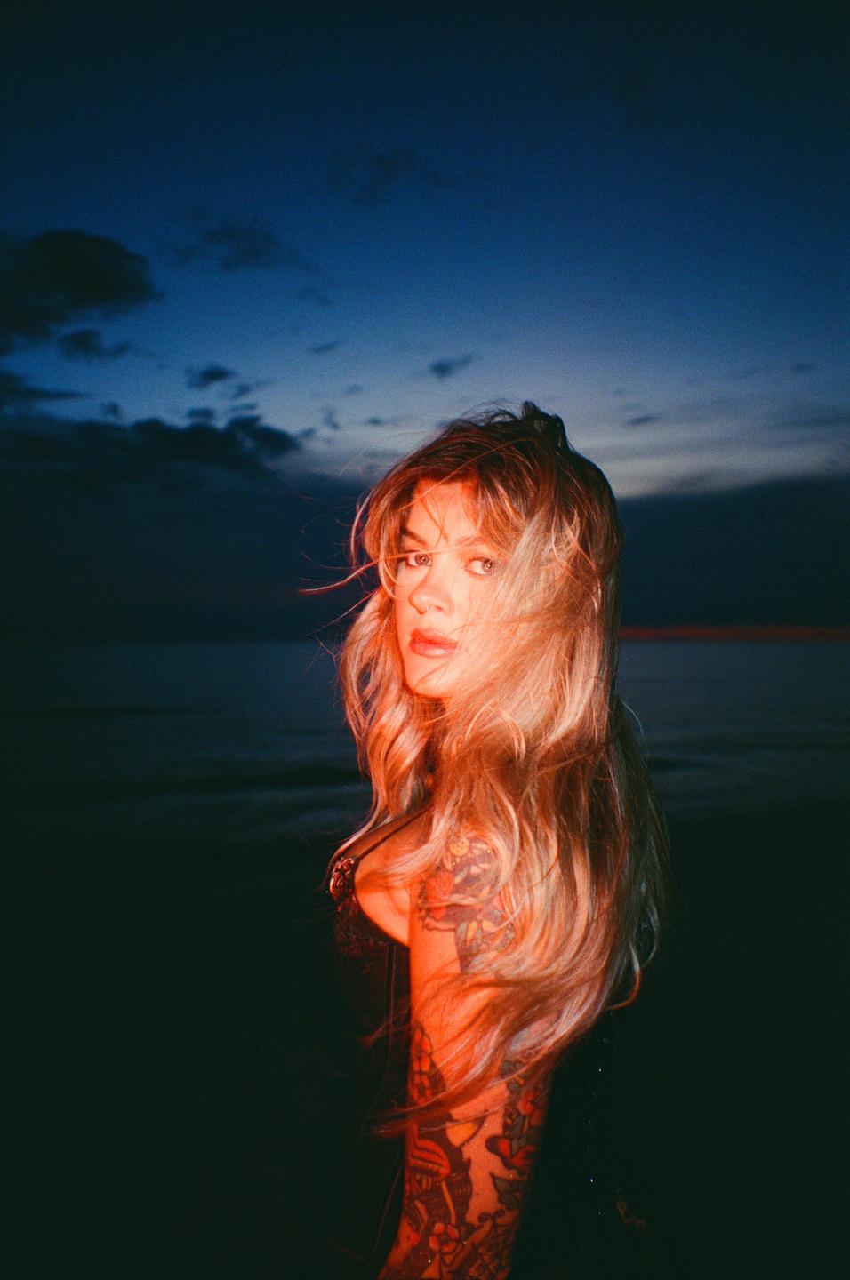 A woman with long blonde hair and tattooed arms looks over her shoulder at dusk.