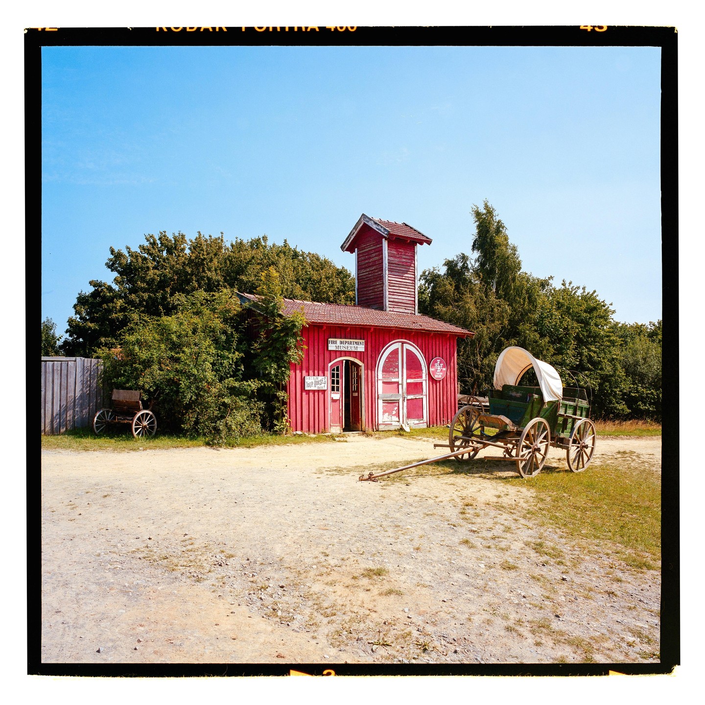 A red fire department museum building with a tower stands next to a covered wagon on a dirt road.