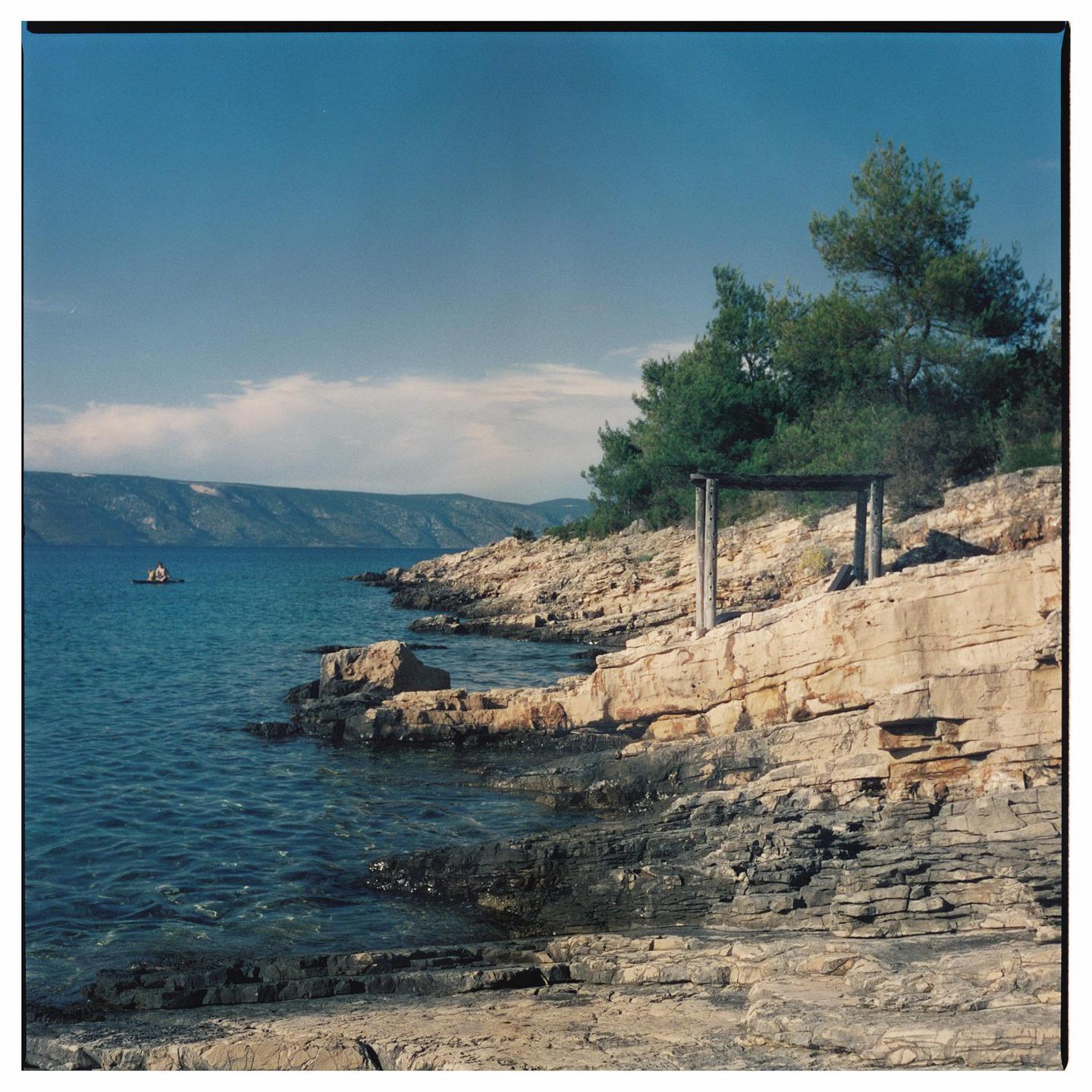 A rocky coastline with a wooden structure and trees on the right, and a small boat with two people in the water on the left.